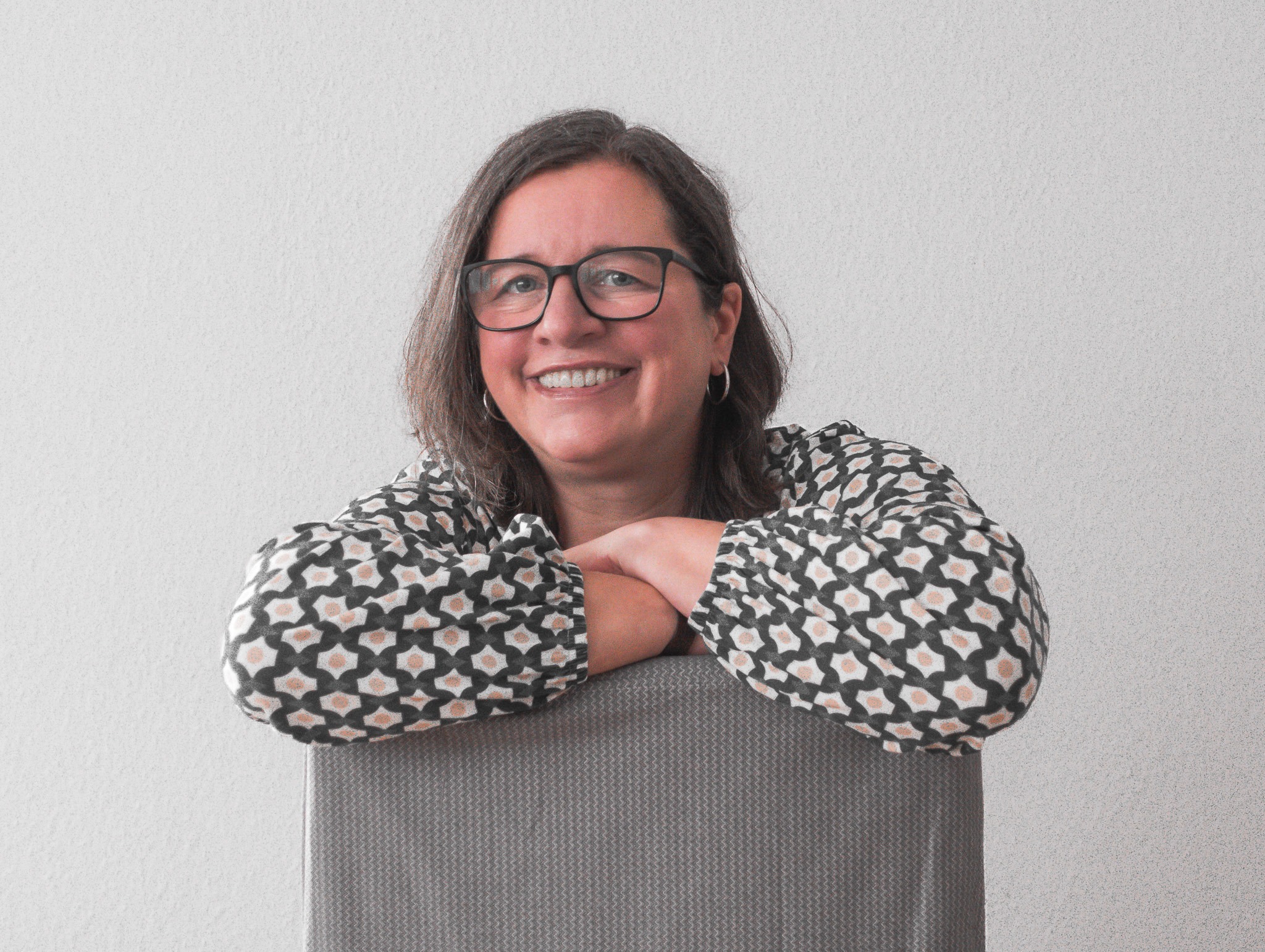 Lächelnde Frau mit Brille lehnt sich entspannt auf eine Stuhllehne.