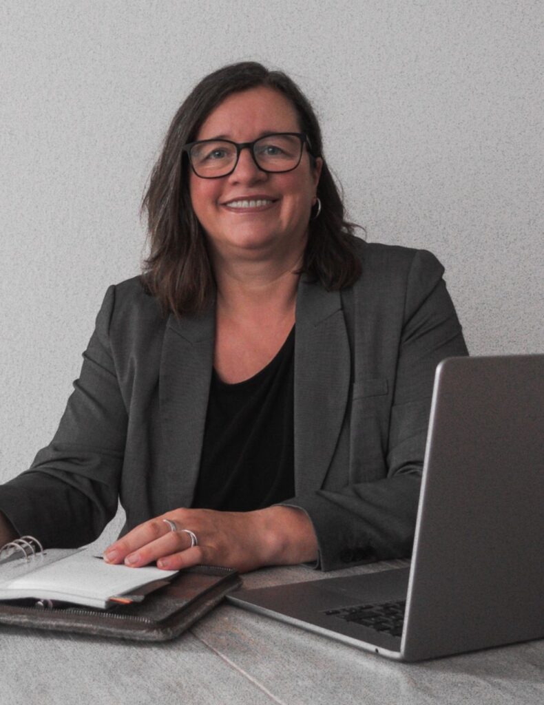 Frau im Business-Outfit schreibt in ein offenes Notizbuch, daneben ein Laptop.