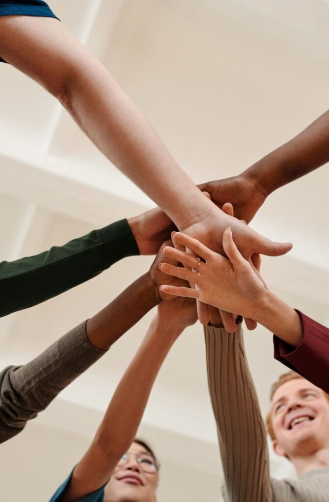 Hände verschiedener Personen legen sich von unten nach oben in einer Teamgeste übereinander.